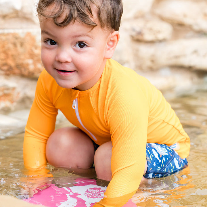 Little one wearing UV protective long sleeve rash top in neon orange. They are also wearing blue jammers.