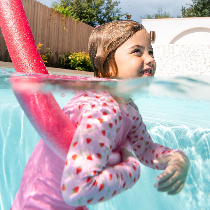 Little one swimming in UV protective long sleeve rash top in pink, and strawberries themed print on the chest and sleeves.