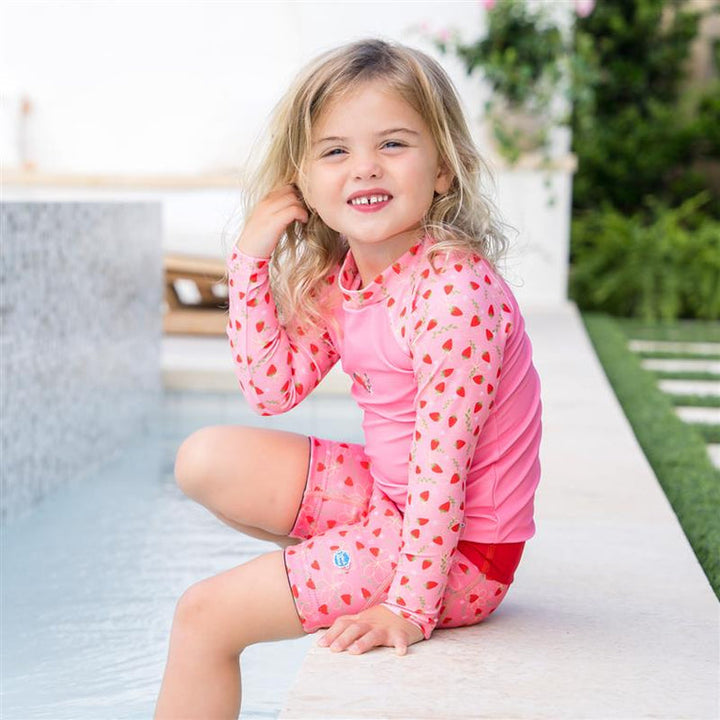 Little one wearing UV protective long sleeve rash top in pink, and strawberries themed print on the chest and sleeves. She is also wearing matching jammers.
