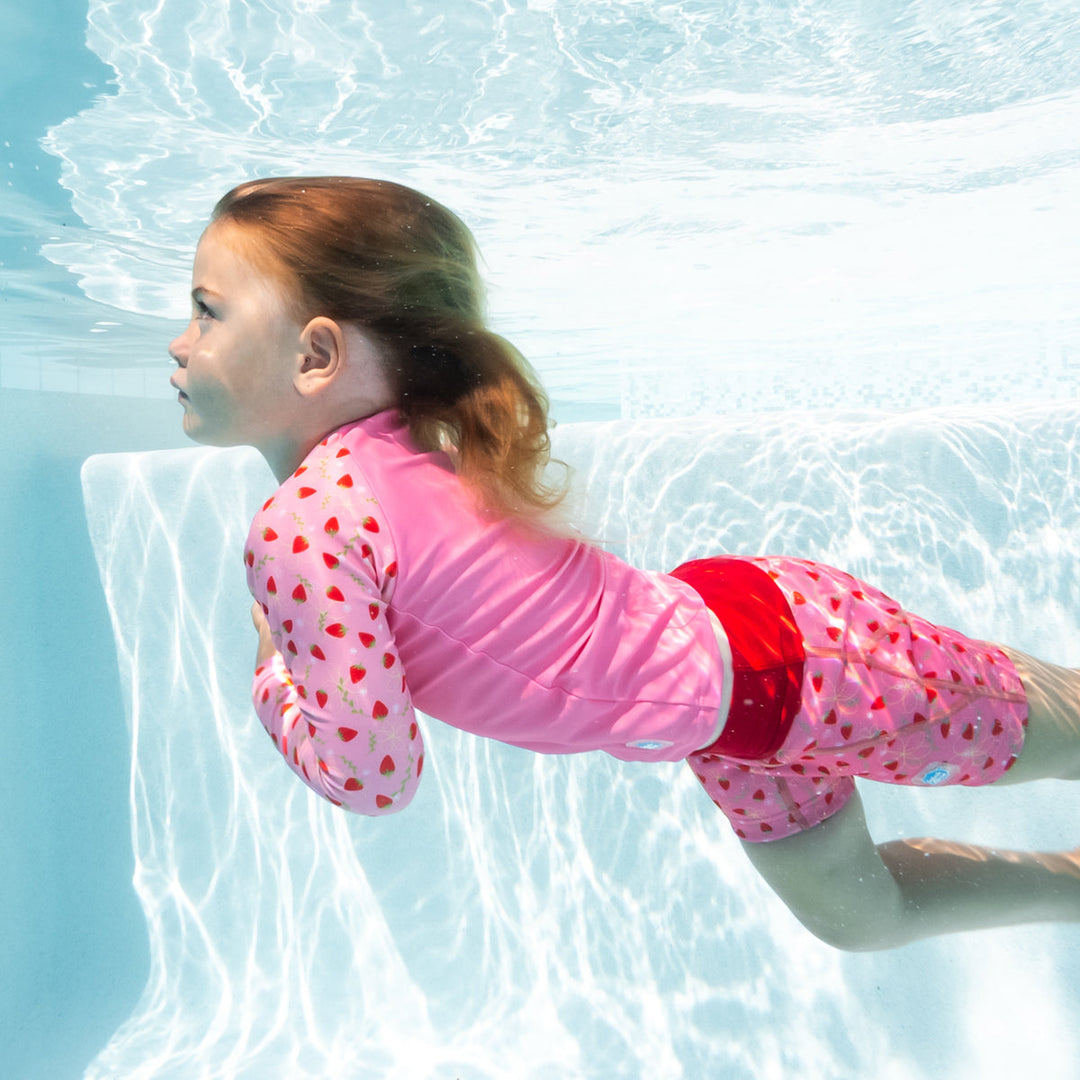 Little one swimming in UV protective long sleeve rash top in pink, and strawberries themed print on the chest and sleeves. She is also wearing matching jammers.