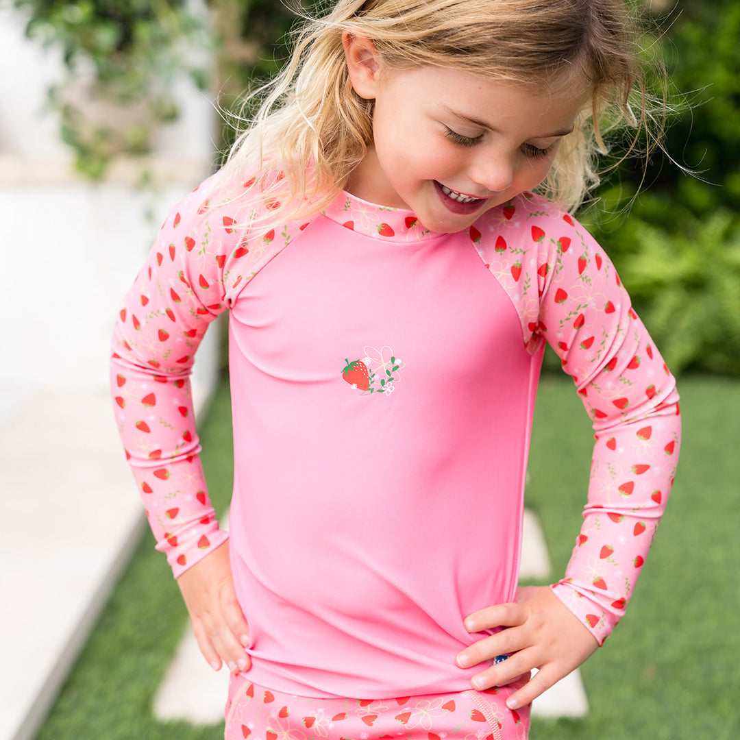 Little one wearing UV protective long sleeve rash top in pink, and strawberries themed print on the chest and sleeves. She is also wearing matching jammers.