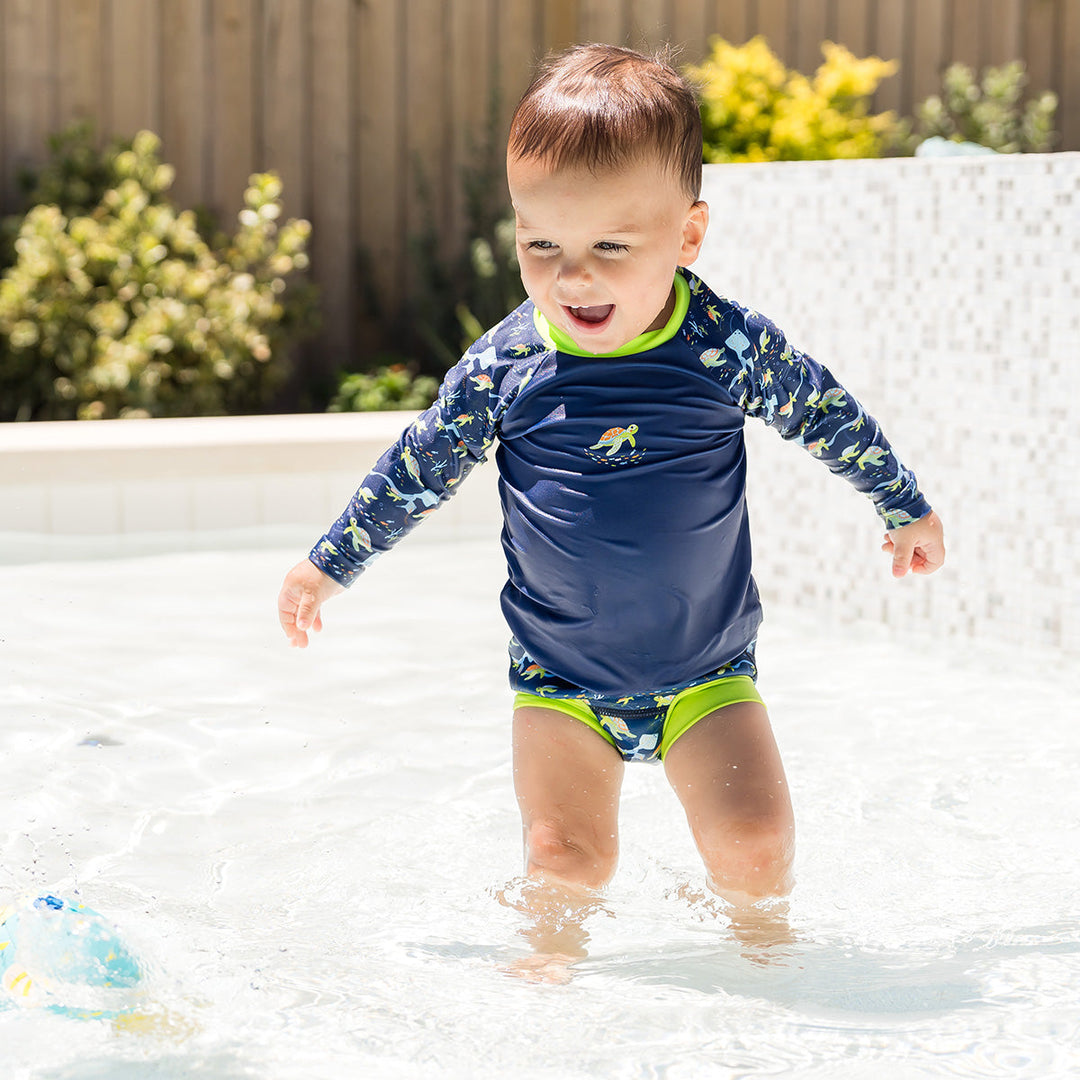 Little one UV protective long sleeve rash top in navy, featuring bright green neckline and crocodiles and frogs themed print on the chest and sleeves. They are wearing matching Happy Nappy.