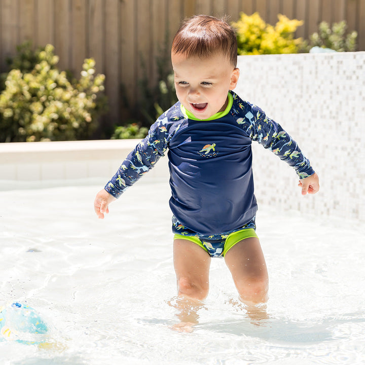 Little one UV protective long sleeve rash top in navy, featuring bright green neckline and crocodiles and frogs themed print on the chest and sleeves. They are wearing matching Happy Nappy.