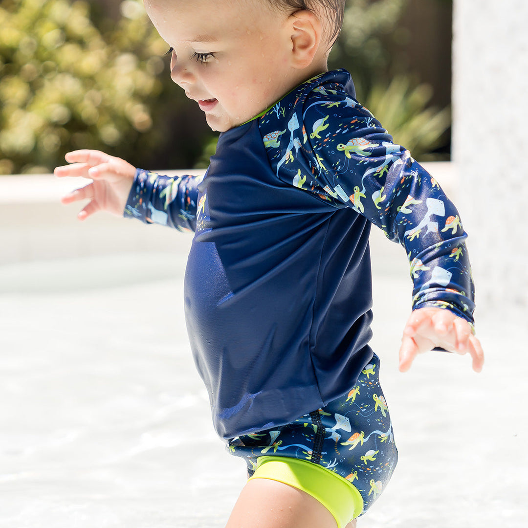 Little one UV protective long sleeve rash top in navy, featuring bright green neckline and crocodiles and frogs themed print on the chest and sleeves. They are wearing matching Happy Nappy.