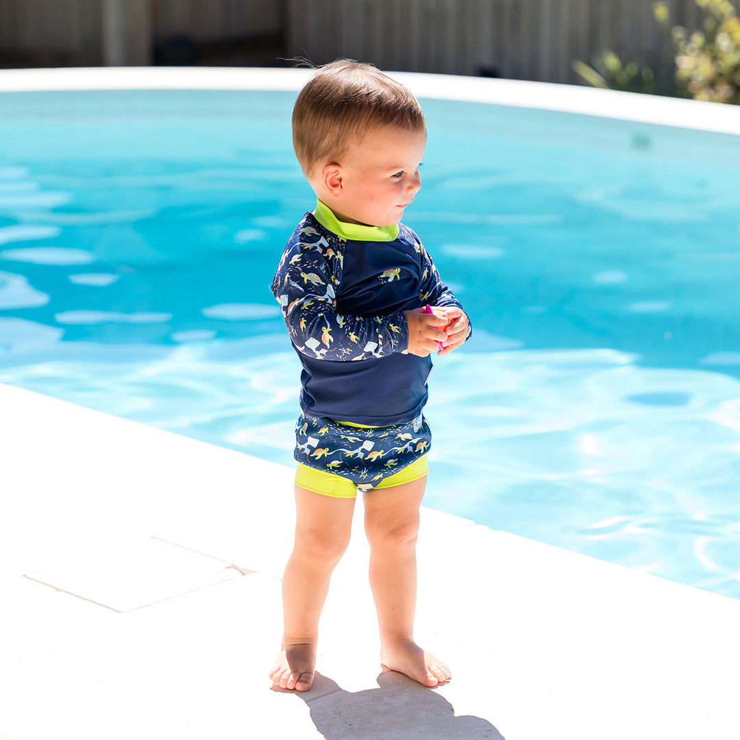 Little one UV protective long sleeve rash top in navy, featuring bright green neckline and crocodiles and frogs themed print on the chest and sleeves. They are wearing matching Happy Nappy.