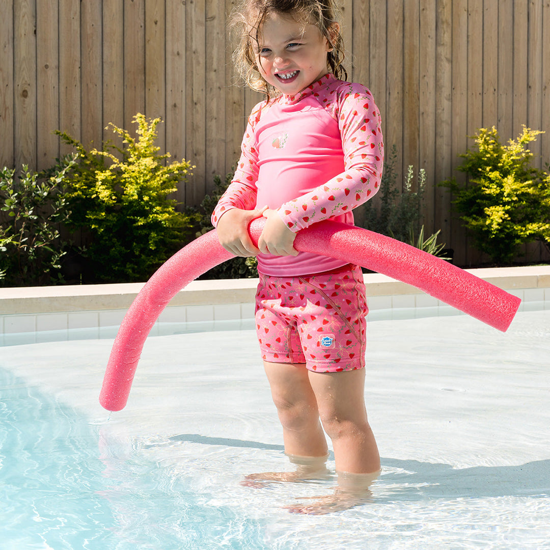 Little one wearing UV protective long sleeve rash top in pink, and strawberries themed print on the chest and sleeves. She is also wearing matching jammers.