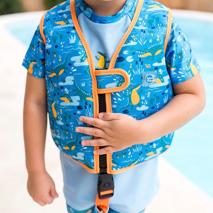 Little one wearing swim vest with non-removable floats in blue, bright orange trims, and crocodiles and frogs themed print. Open.