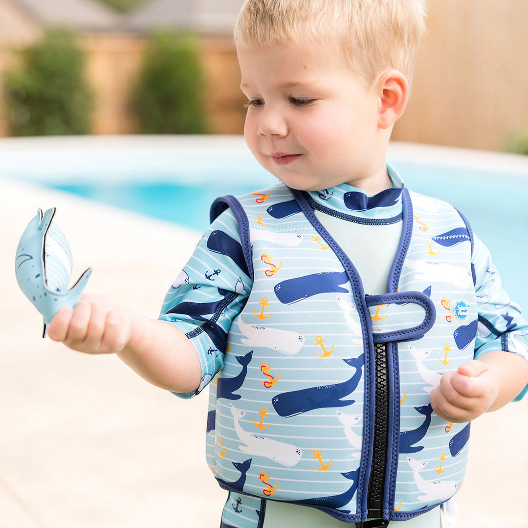 Lifestyle image of child wearing neoprene swim vest with non-removable floats in light blue, navy trims, and whales and anchors themed print. Front.