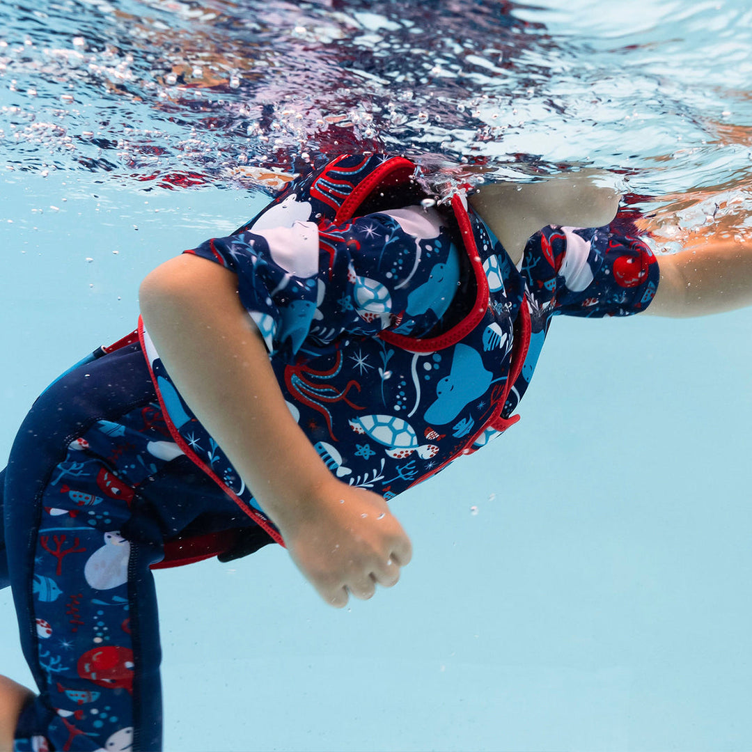 Lifestyle image of child swimming with neoprene swim vest with non-removable floats in navy, red trims, and sea life themed print. Side.