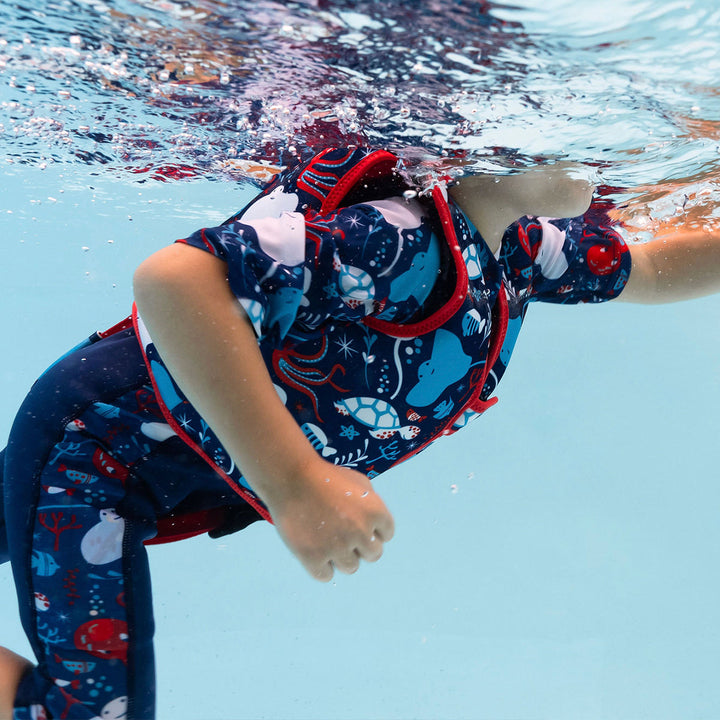 Lifestyle image of child swimming with neoprene swim vest with non-removable floats in navy, red trims, and sea life themed print. Side.