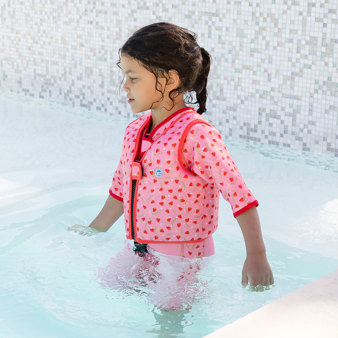 Lifestyle image of child wearing neoprene swim vest with non-removable floats in pink, red trims and strawberries themed print. Side.