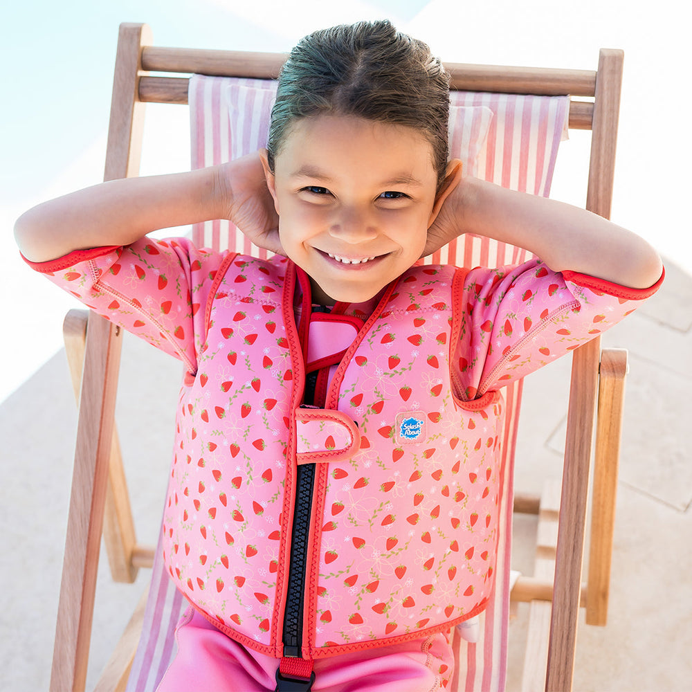 Lifestyle image of child wearing neoprene swim vest with non-removable floats in pink, red trims and strawberries themed print. Front.