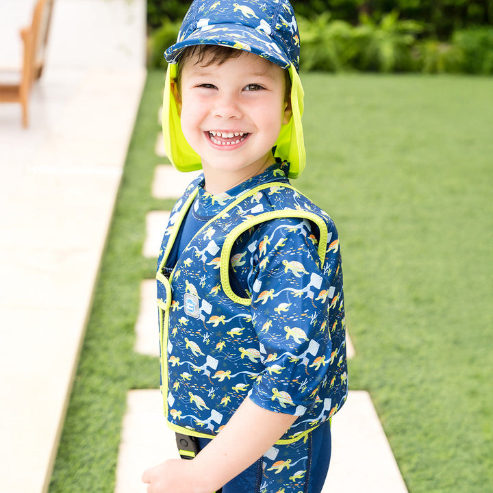 Little one wearing swim vest for toddlers with non-removable floats in navy, bright green trims, and turtles and mantarays themed print. He's wearing matching legionnaire hat.