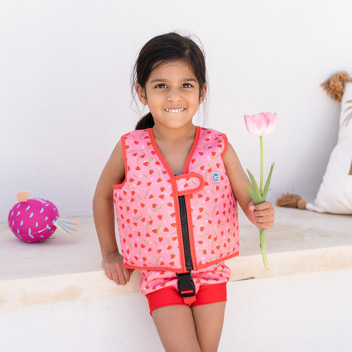 Lifestyle image of child wearing neoprene swim vest with non-removable floats in pink, red trims and strawberries themed print. Front.