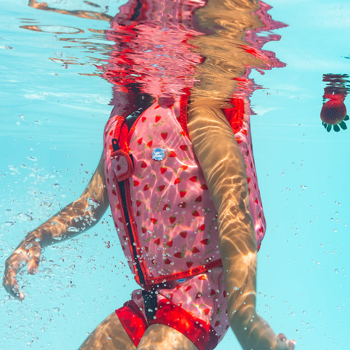 Lifestyle image of child wearing neoprene swim vest with non-removable floats in pink, red trims and strawberries themed print. Side.