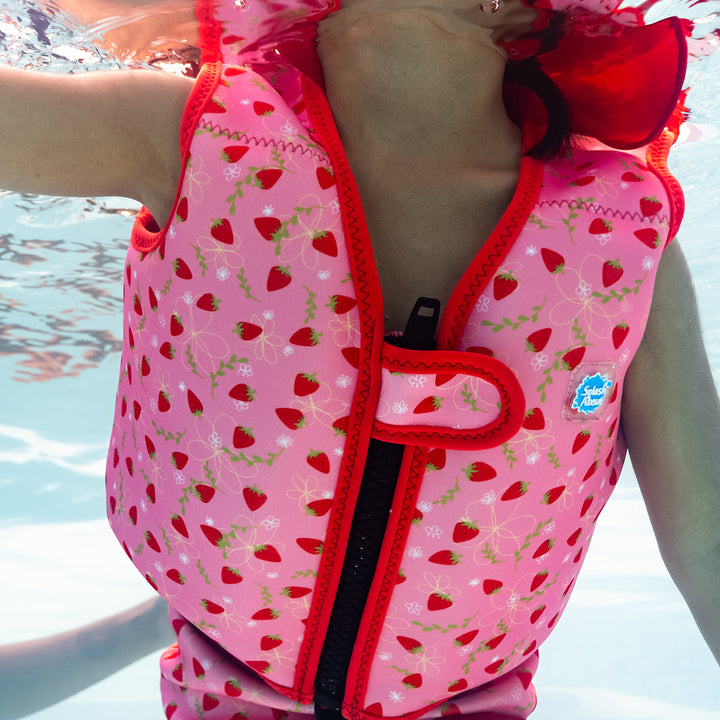 Lifestyle image of child wearing neoprene swim vest with non-removable floats in pink, red trims and strawberries themed print. Front.