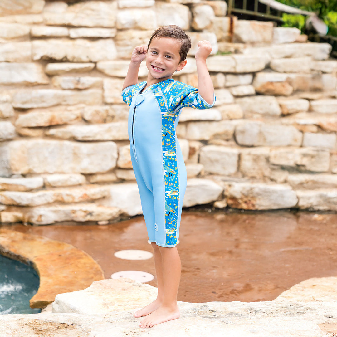Kid wearing shorty wetsuit in light blue featuring crocodiles and frogs themed print in sleeves and side panels.