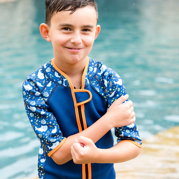Kid wearing shorty wetsuit in navy with bright orange trims, featuring sharks and coral themed print in sleeves and side panels.