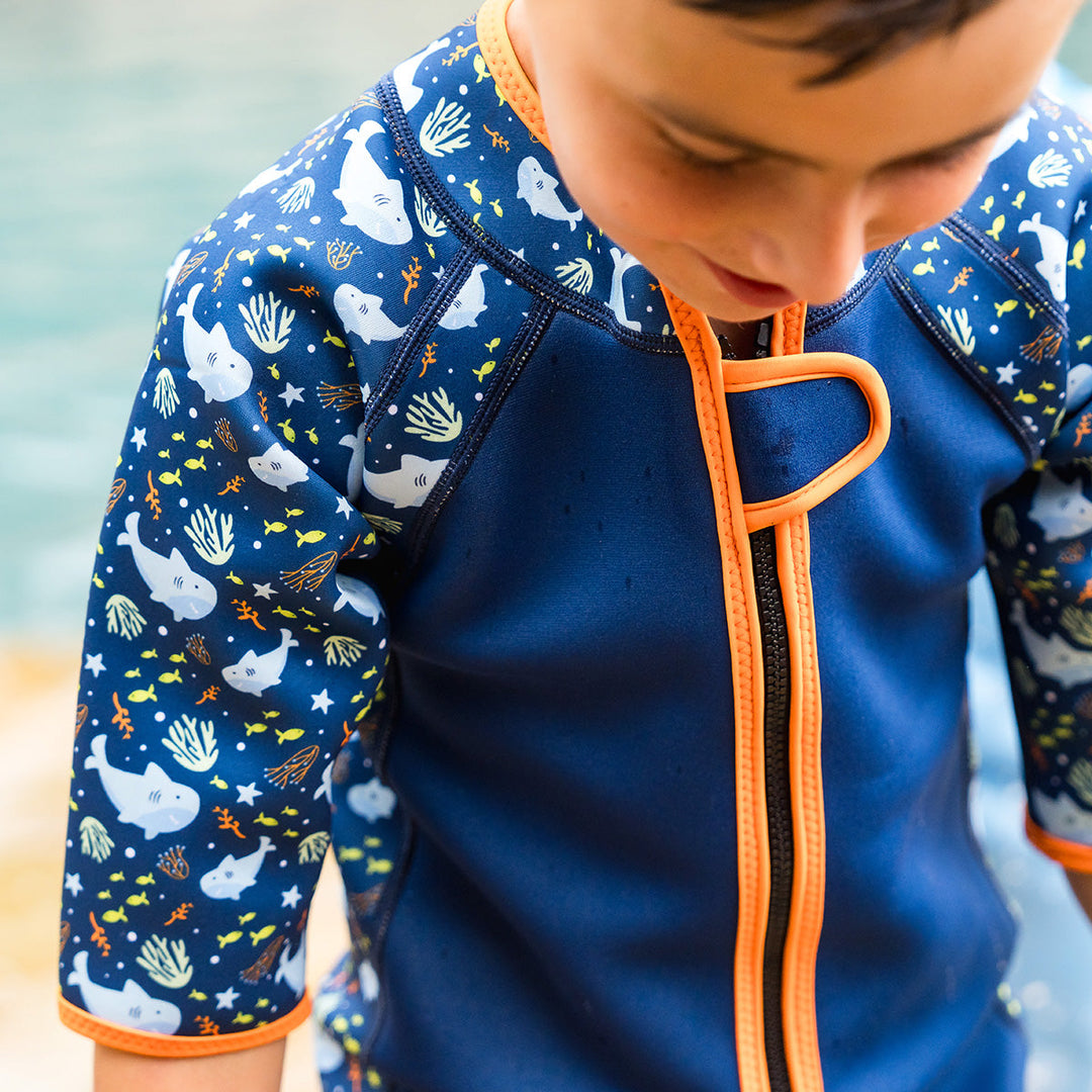 Close-up of front zip of shorty wetsuit in navy with bright orange trims, featuring sharks and coral themed print in sleeves and side panels.
