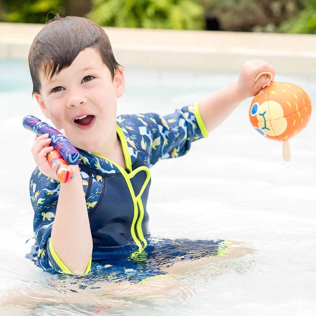 Kid swimming in a shorty wetsuit in navy with bright green trims, featuring turtles and mantarays themed print in sleeves and side panels.