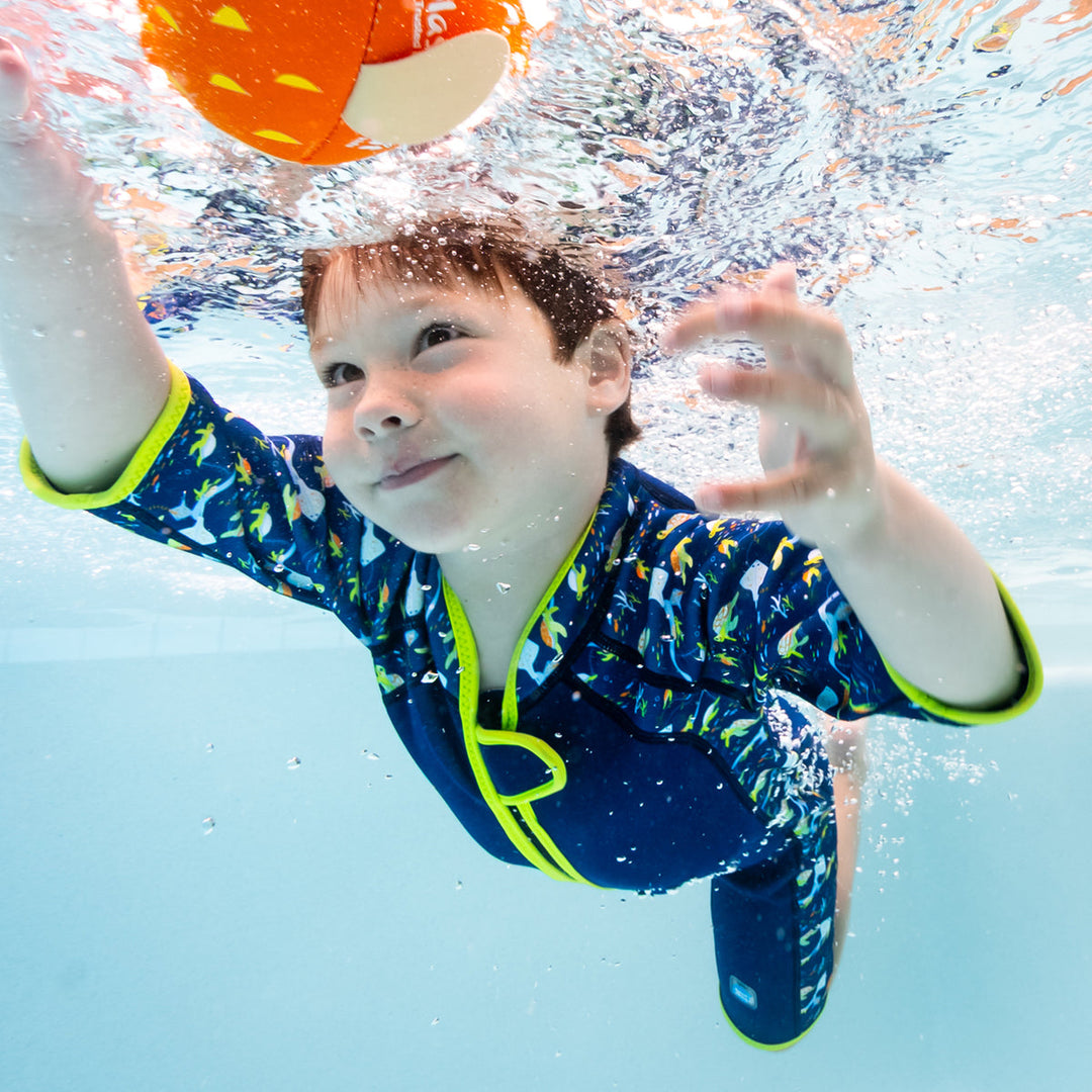 Kid swimming in a shorty wetsuit in navy with bright green trims, featuring turtles and mantarays themed print in sleeves and side panels.