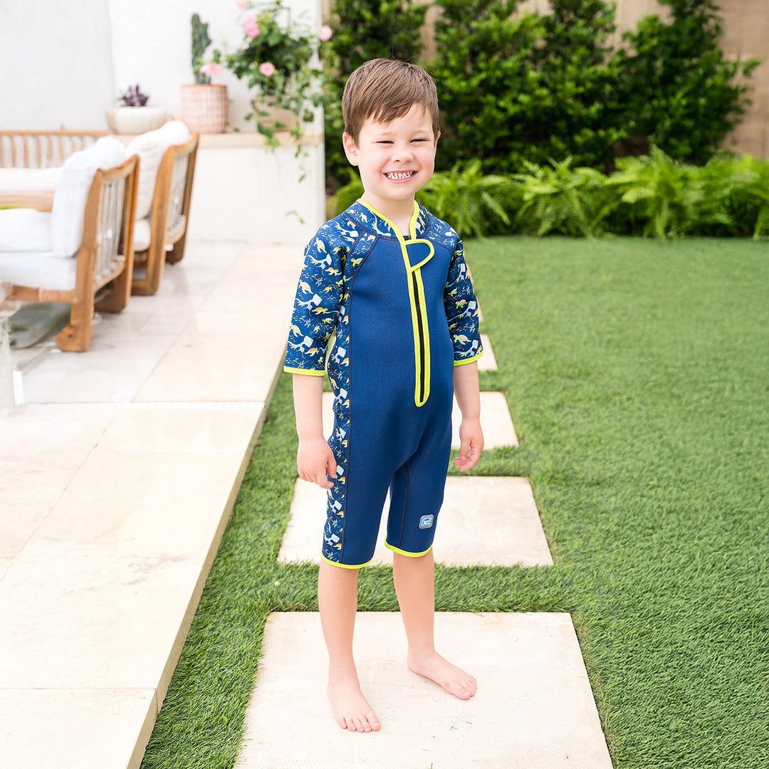 Kid wearing a shorty wetsuit in navy with bright green trims, featuring turtles and mantarays themed print in sleeves and side panels.