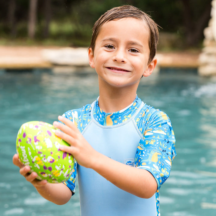 Child wearing one piece UV sun and sea wetsuit for toddlers in blue, featuring crocodiles and frogs themed print on sleeves, side panels, neck and chest. Back.
