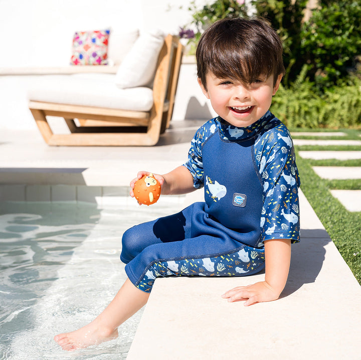 Child wearing one piece UV sun and sea wetsuit for toddlers in navy blue, featuring sharks, coral and fish themed print on sleeves, side panels, neck and chest. Poolside.