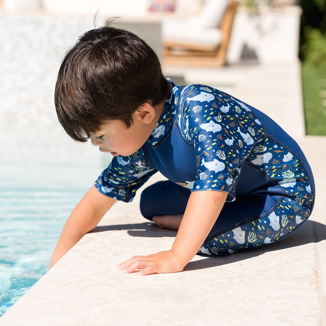 Child wearing one piece UV sun and sea wetsuit for toddlers in navy blue, featuring sharks, coral and fish themed print on sleeves, side panels, neck and chest. Poolside.