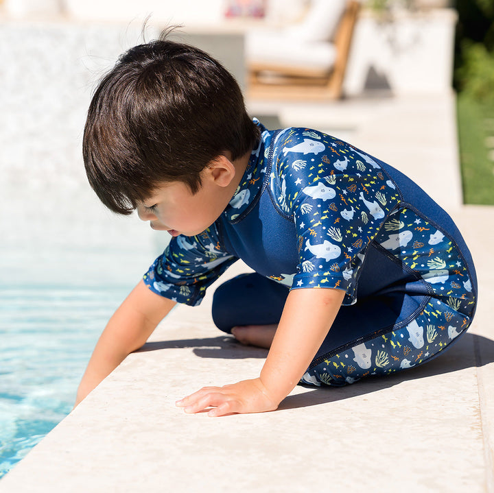 Child wearing one piece UV sun and sea wetsuit for toddlers in navy blue, featuring sharks, coral and fish themed print on sleeves, side panels, neck and chest. Poolside.