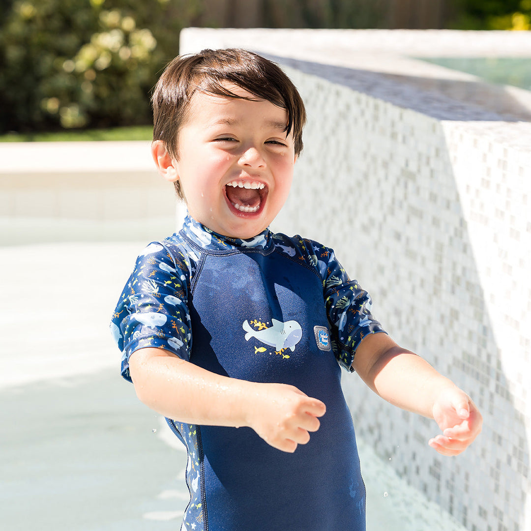 Child wearing one piece UV sun and sea wetsuit for toddlers in navy blue, featuring sharks, coral and fish themed print on sleeves, side panels, neck and chest.