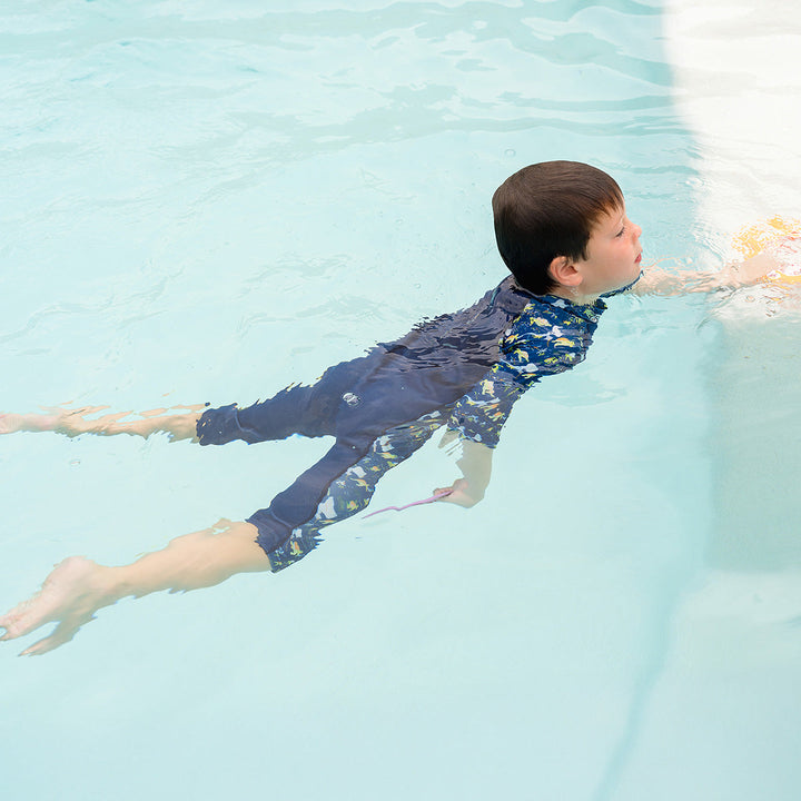 Child swimming in one piece UV sun and sea wetsuit for toddlers in navy blue, featuring sea turtles, coral and fish themed print on sleeves, side panels, neck and chest.