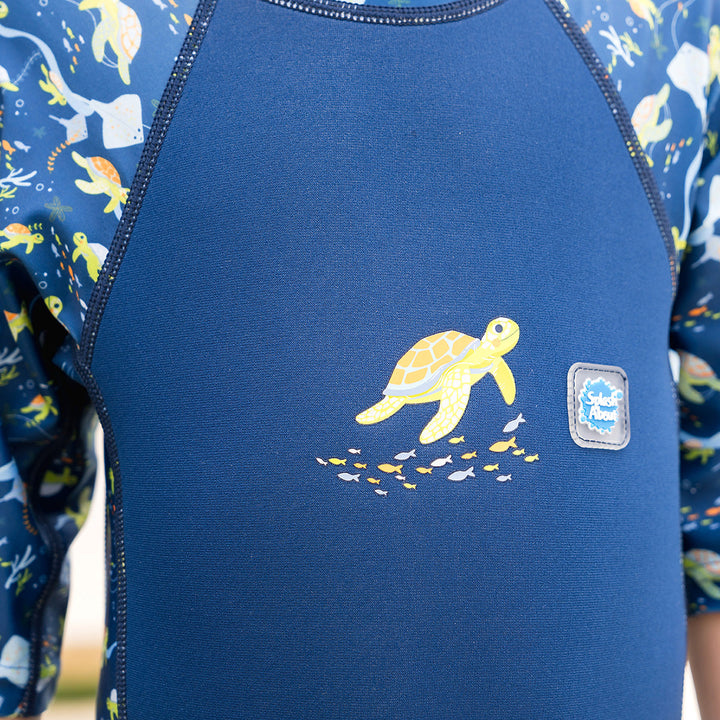 Close-up of child wearing one piece UV sun and sea wetsuit for toddlers in navy blue, featuring sea turtles, coral and fish themed print on sleeves, side panels, neck and chest.