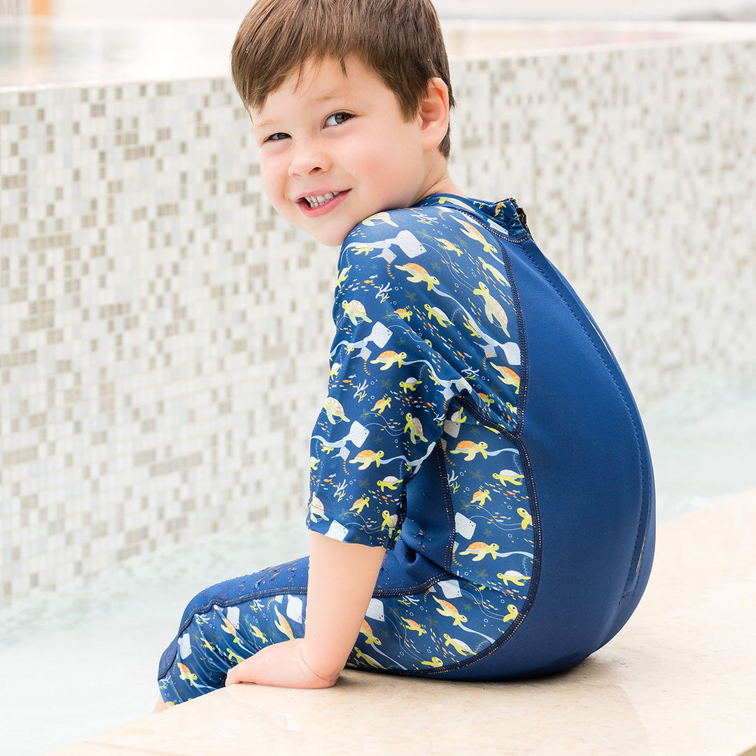 Child wearing one piece UV sun and sea wetsuit for toddlers in navy blue, featuring sea turtles, coral and fish themed print on sleeves, side panels, neck and chest. Poolside.