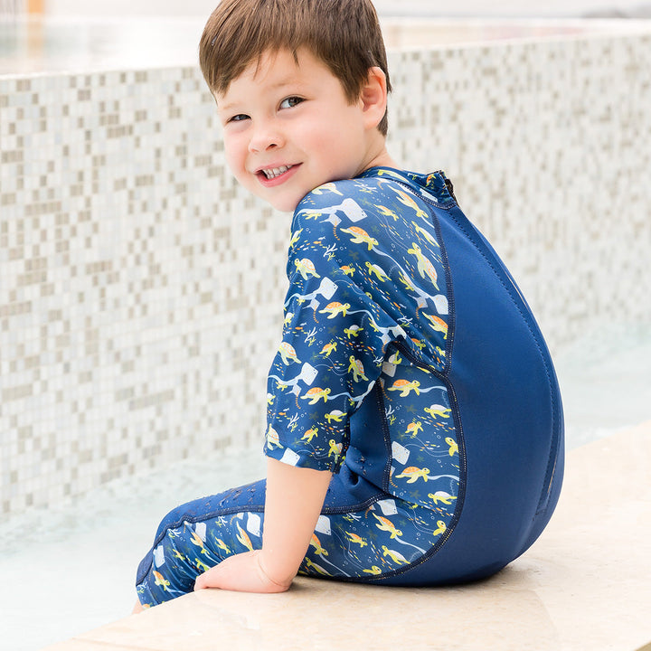 Child wearing one piece UV sun and sea wetsuit for toddlers in navy blue, featuring sea turtles, coral and fish themed print on sleeves, side panels, neck and chest. Poolside.