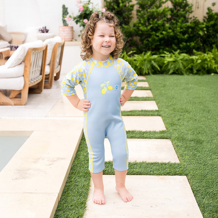 Child wearing one piece UV sun and sea wetsuit for toddlers in light blue, featuring lemons themed print on sleeves, side panels, neck and chest.