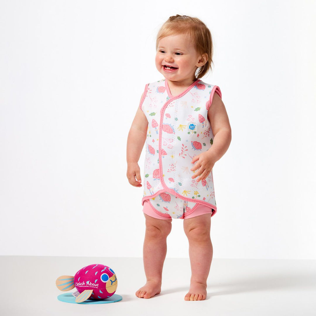 Little one wearing a neoprene baby wrap wetsuit in white with pink trims and forest themed print, including hedgehogs, mushrooms, flowers and leaves. Side.