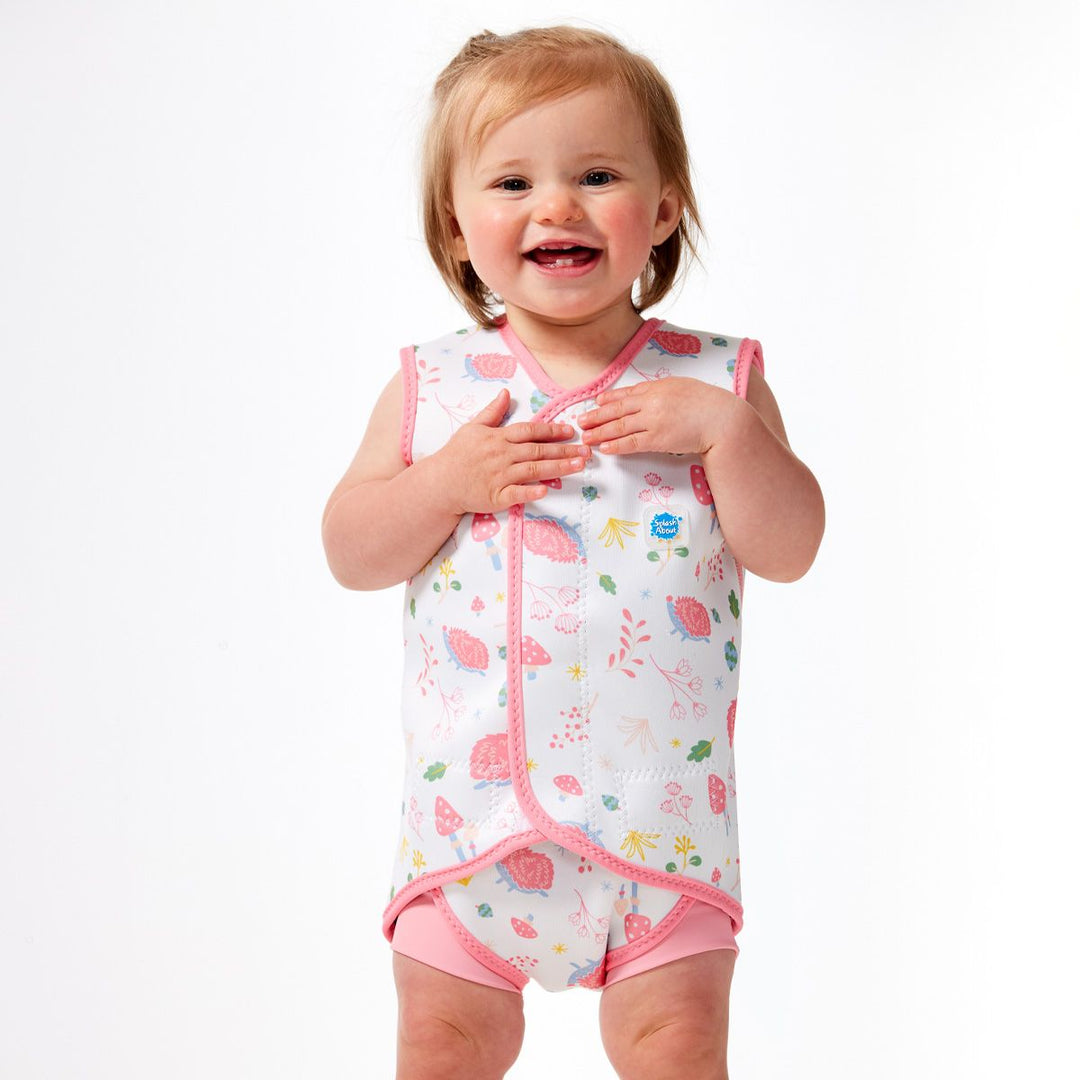 Little one wearing a neoprene baby wrap wetsuit in white with pink trims and forest themed print, including hedgehogs, mushrooms, flowers and leaves. Front.