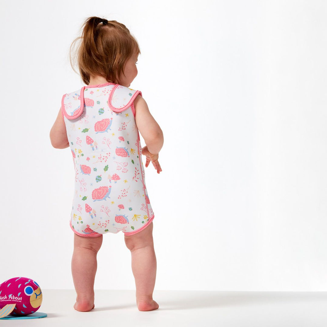 Little one wearing a neoprene baby wrap wetsuit in white with pink trims and forest themed print, including hedgehogs, mushrooms, flowers and leaves. Back.