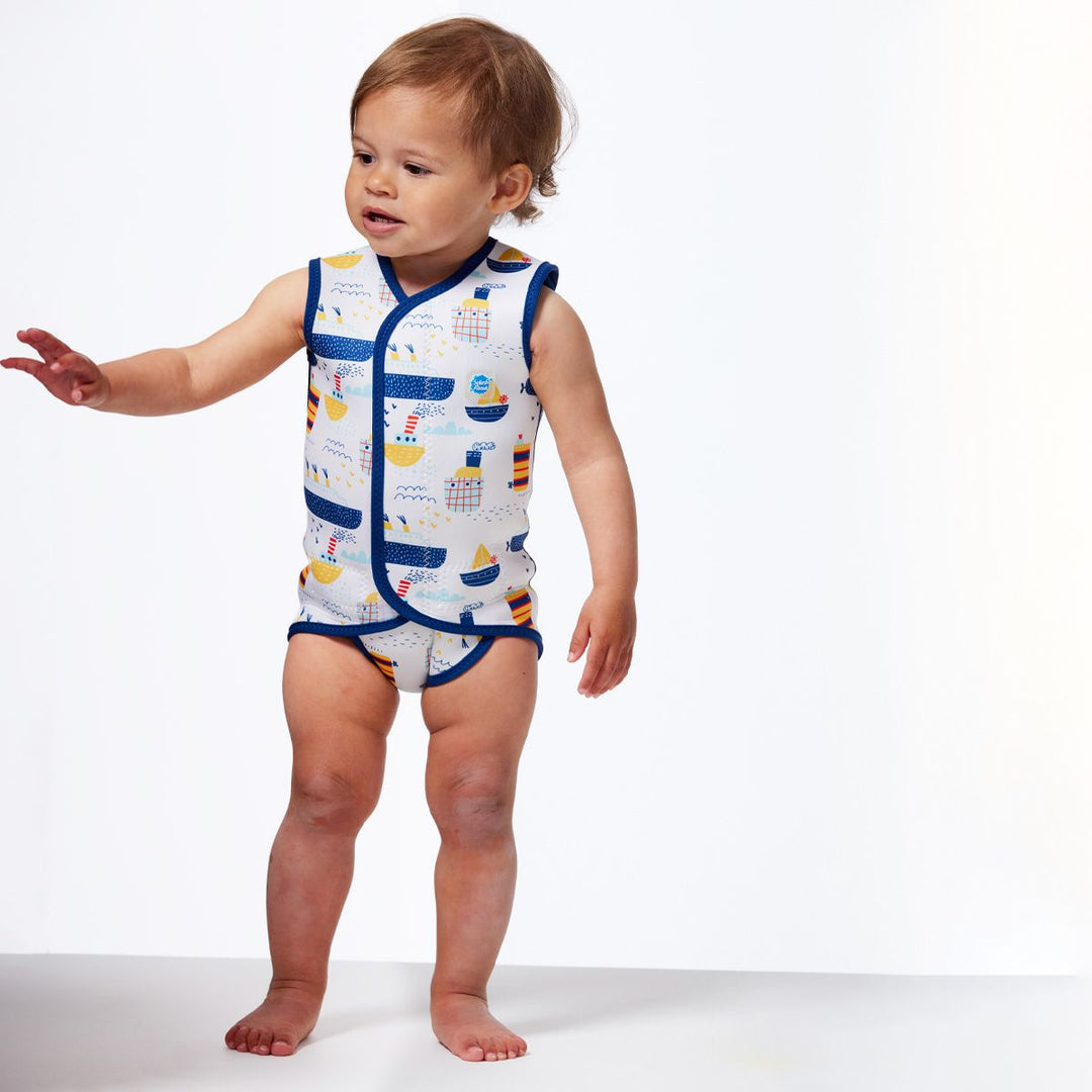 Little one wearing a neoprene baby wrap wetsuit in white with navy trims and boats themed print. Front.