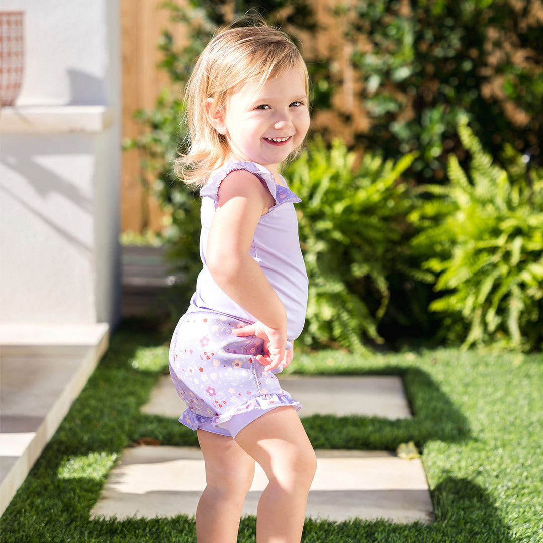 Child wearing Happy Nappy Costume in lilac and floral themed print, featuring frills in arms and legs.