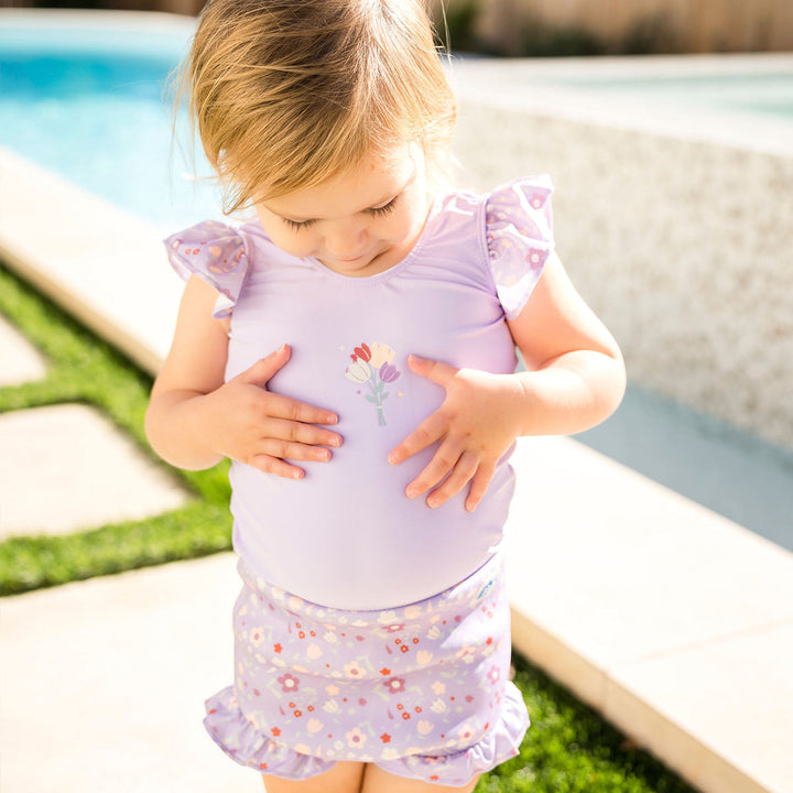 Child wearing Happy Nappy Costume in lilac and floral themed print, featuring frills in arms and legs.