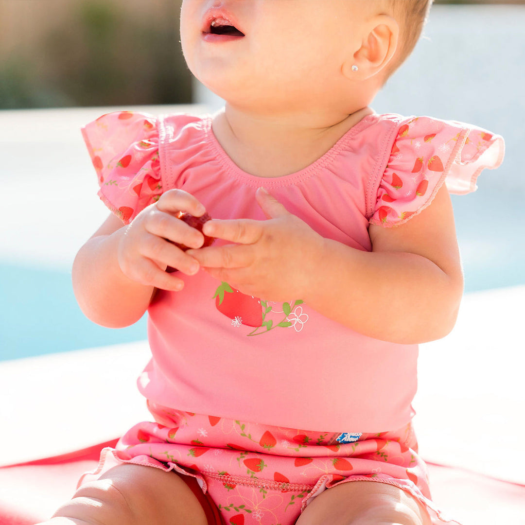 Close up of child wearing Happy Nappy Costume in pink with red trims and strawberries themed print, featuring frills in arms and legs. Front.