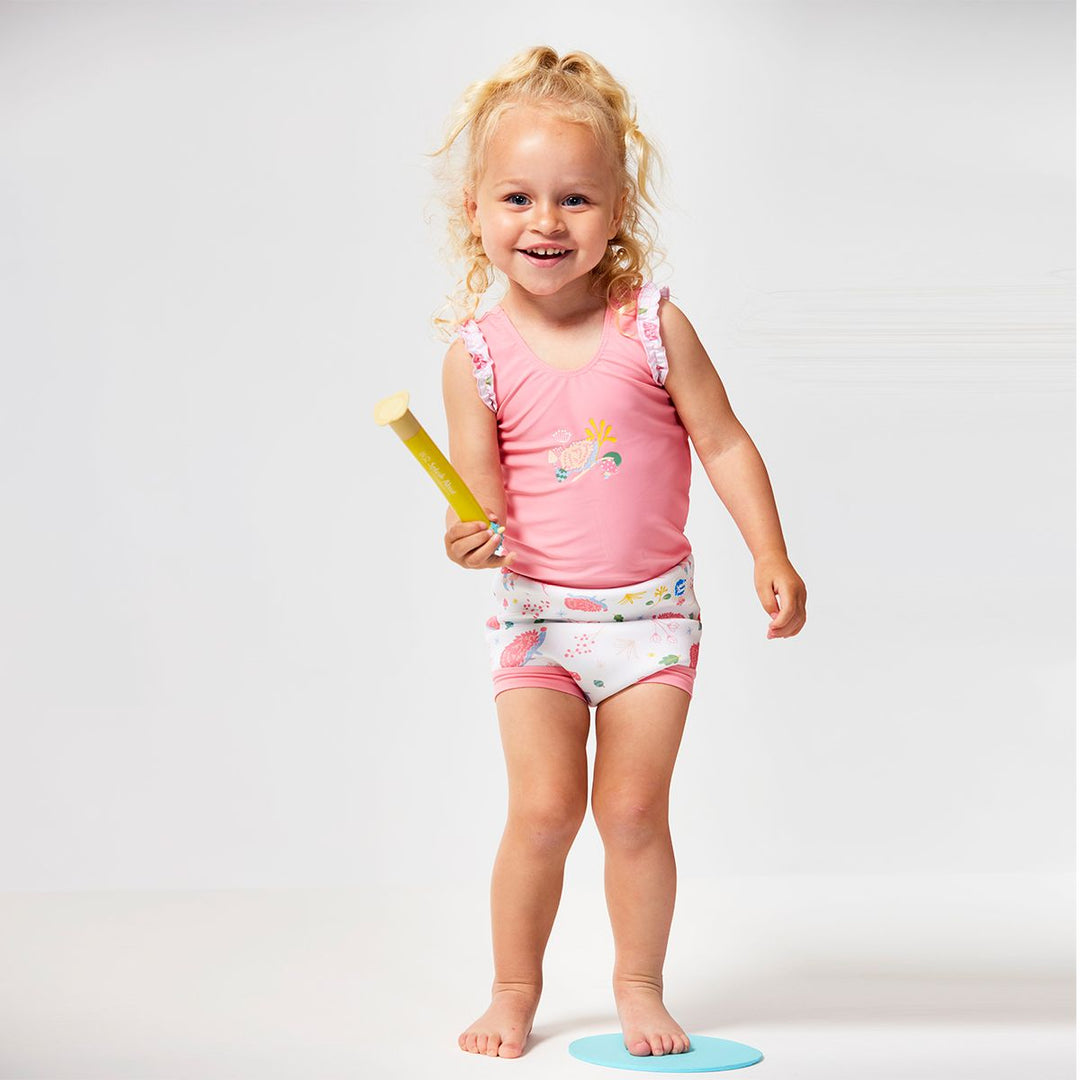 Lifestyle image of child wearing a Happy Nappy Costume in pink and white featuring forest themed print, including hedgehogs, mushrooms and leaves. Front.