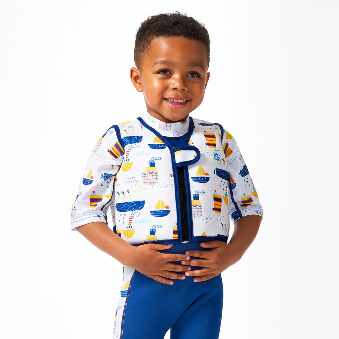 Lifestyle image of child wearing neoprene swim vest for toddlers with non-removable floats in white, navy trims and boats themed print. He's also wearing matching Sun & Sea Wetsuit. Front.