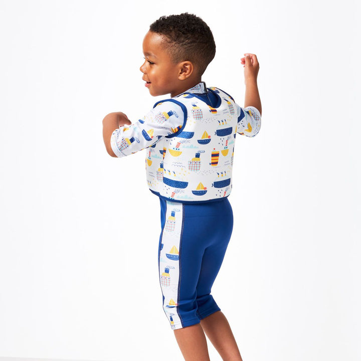 Lifestyle image of child wearing neoprene swim vest for toddlers with non-removable floats in white, navy trims and boats themed print. He's also wearing matching Sun & Sea Wetsuit. Back.