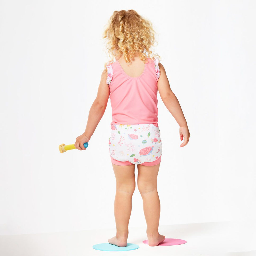 Lifestyle image of child wearing a Happy Nappy Costume in pink and white featuring forest themed print, including hedgehogs, mushrooms and leaves. Back.