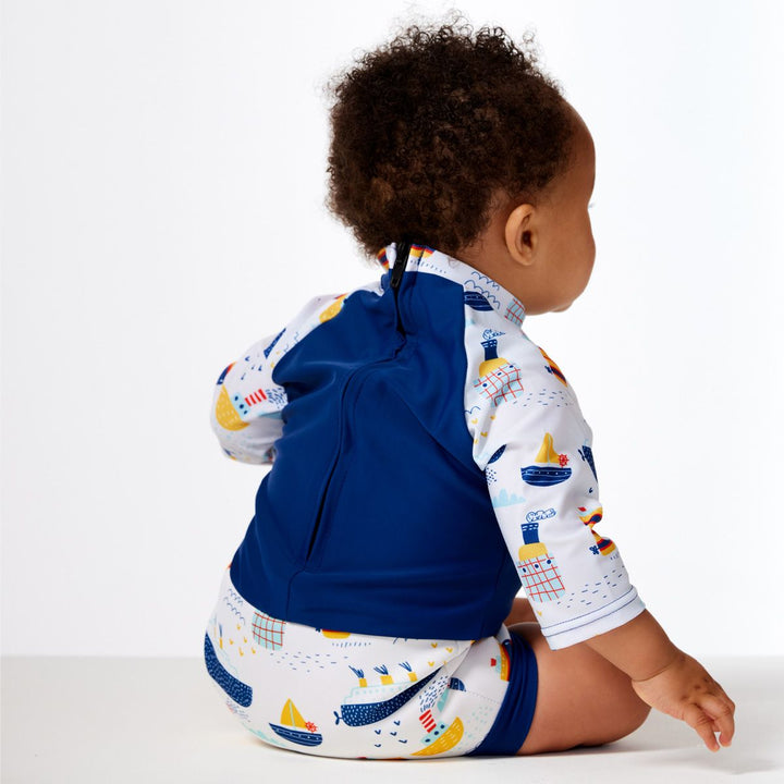Lifestyle image of child wearing Happy Nappy Sunsuit in white and navy, and boats themed print. Back.