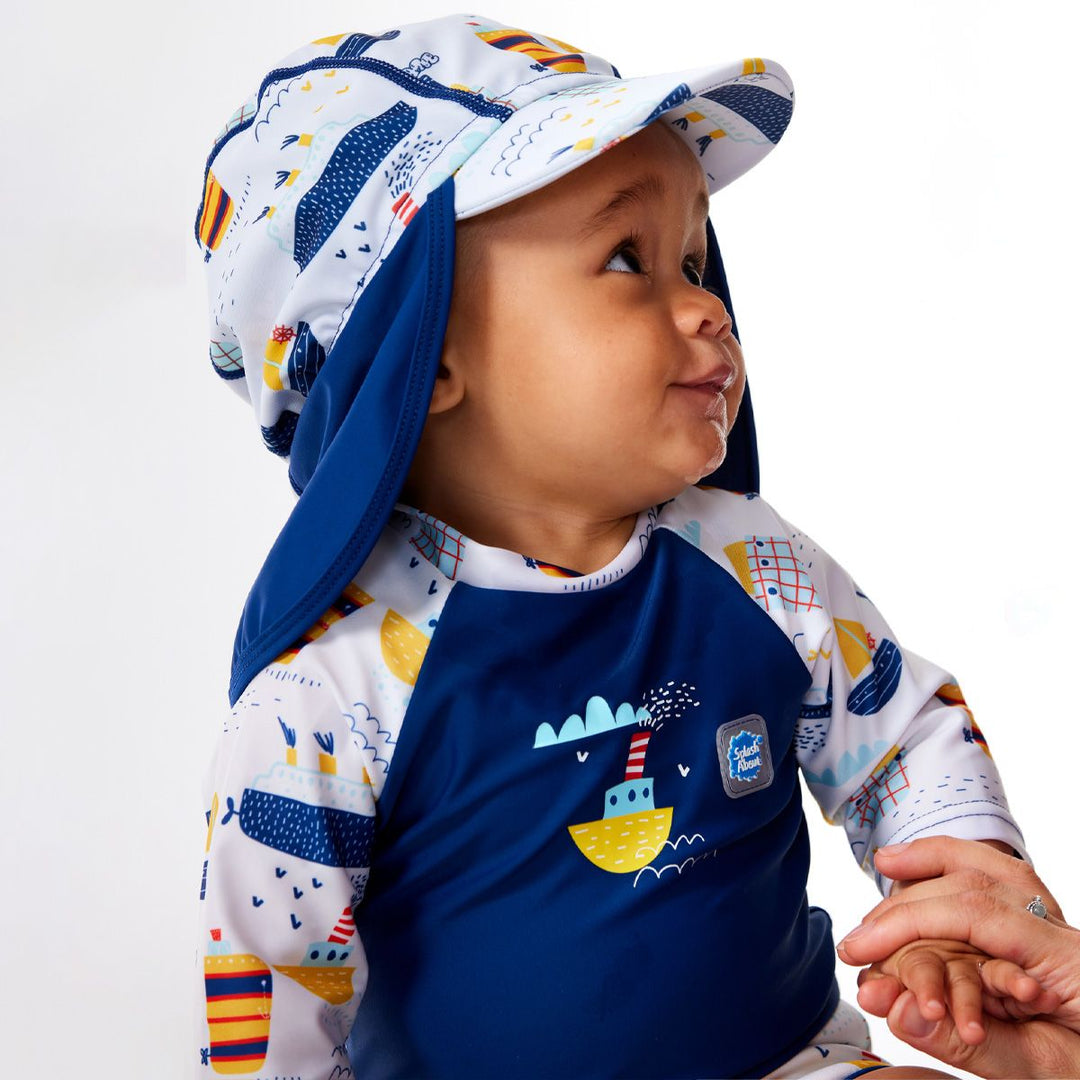 Lifestyle image of child wearing Happy Nappy Sunsuit in white and navy, and boats themed print. They're also wearing matching legionnaire hat. Close-up.