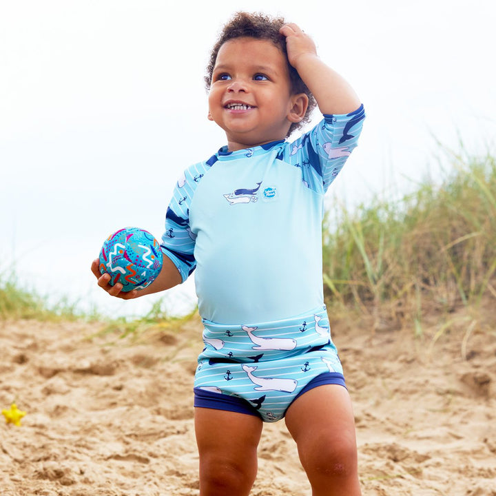 Lifestyle image of baby wearing a Happy Nappy Sunsuit in light blue and whales themed print. Front.